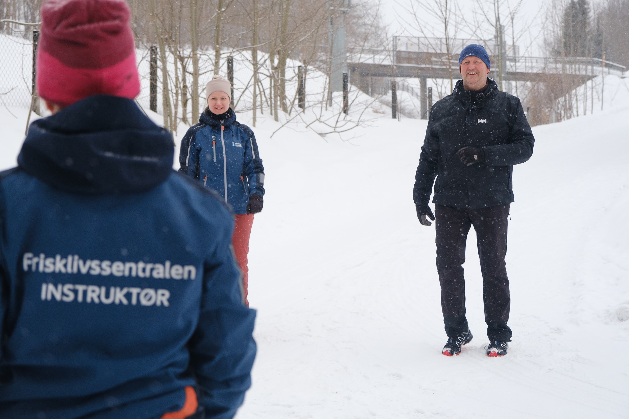 En mann trener ute i snøen. I forgrunn er det en kvinnelig instruktør med ryggen i mot. På ryggen står det frisklivssentralen