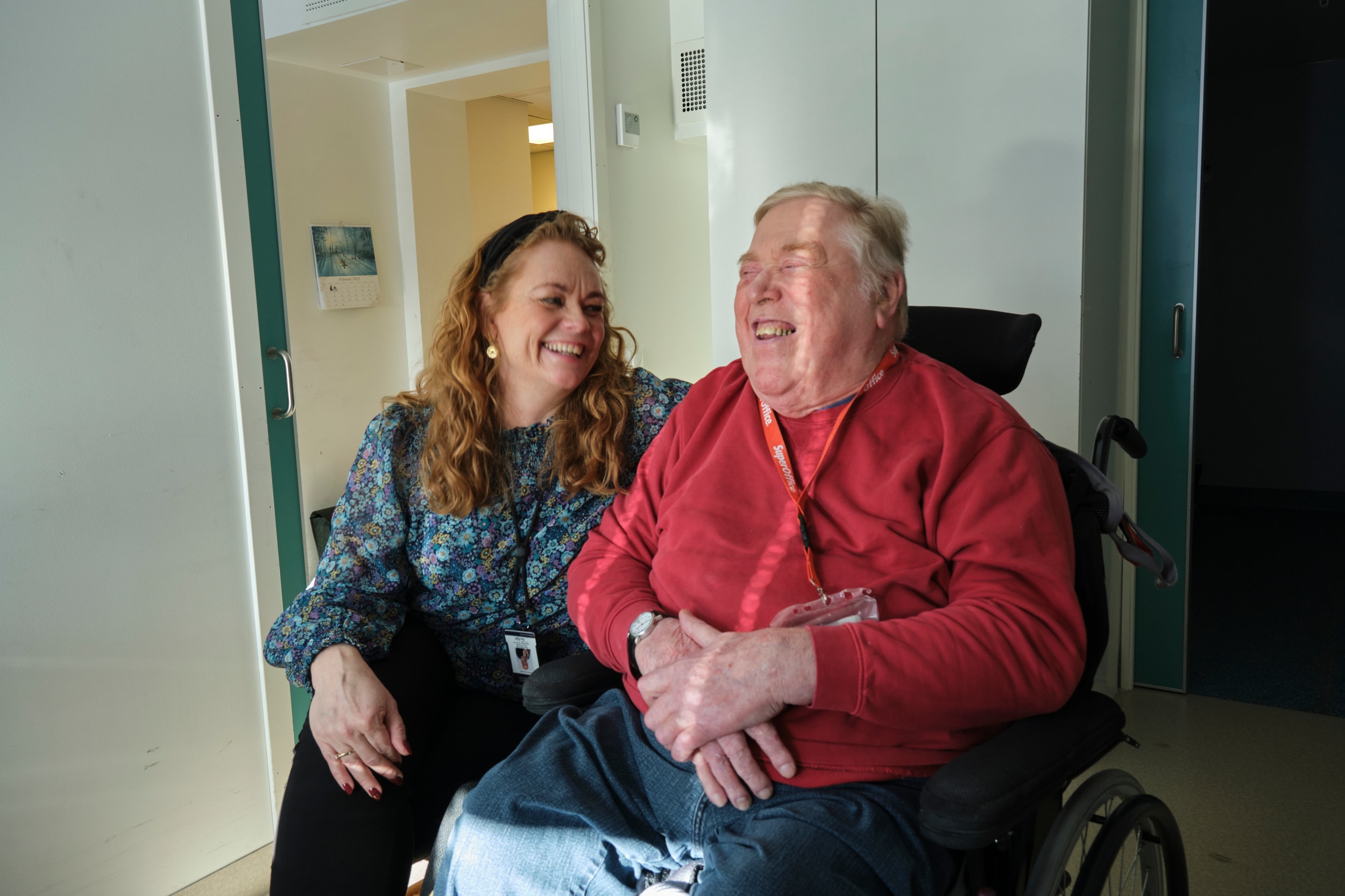 En gammel mann i rullestol med rød genser prater med dame. Begge ler.