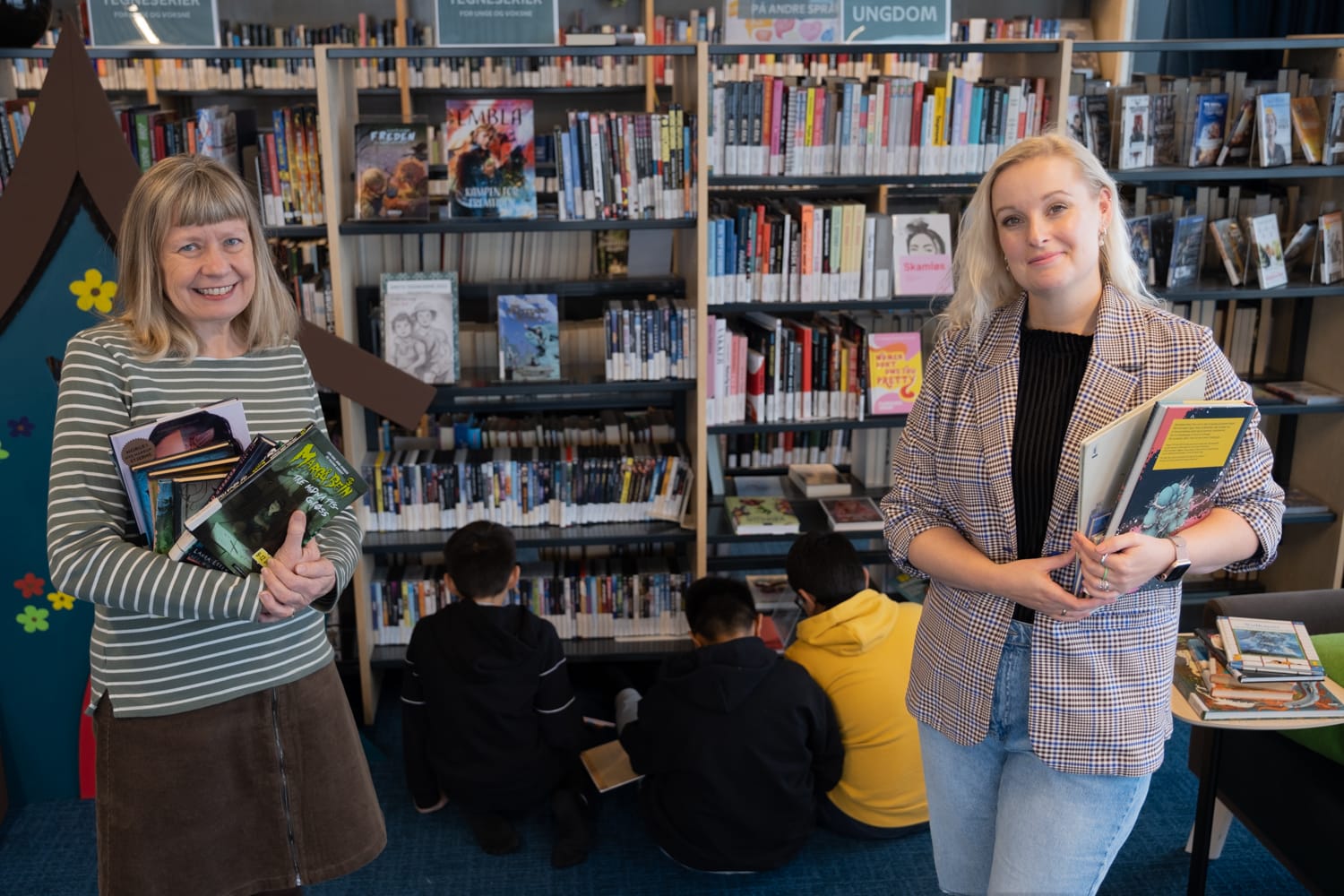 Inspirasjonsbibliotekar Inger Bergan Mortensen og prosjektleder i Drammen kommune, Katrine Krikeby Arnold står foran en bokhylle. Tre barn sitter med ryggen til på gulvet og lseer i bøker. 
