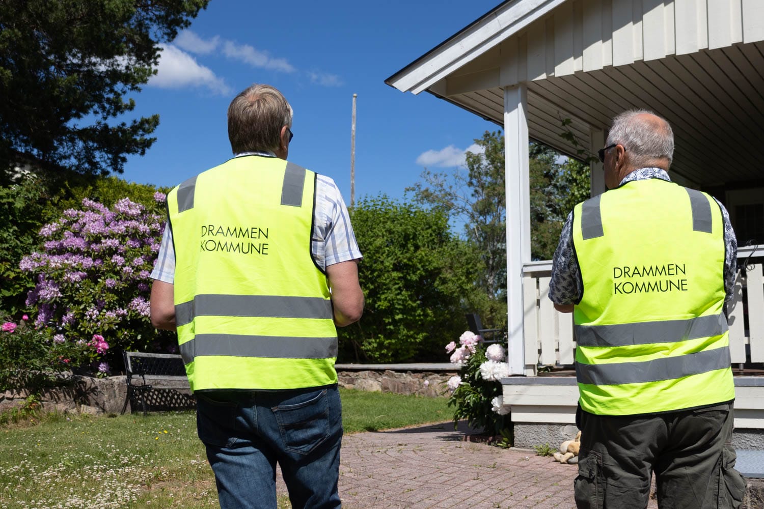 To menn med gul vest går. De er fra Drammen kommune. Foran hvitt hus.