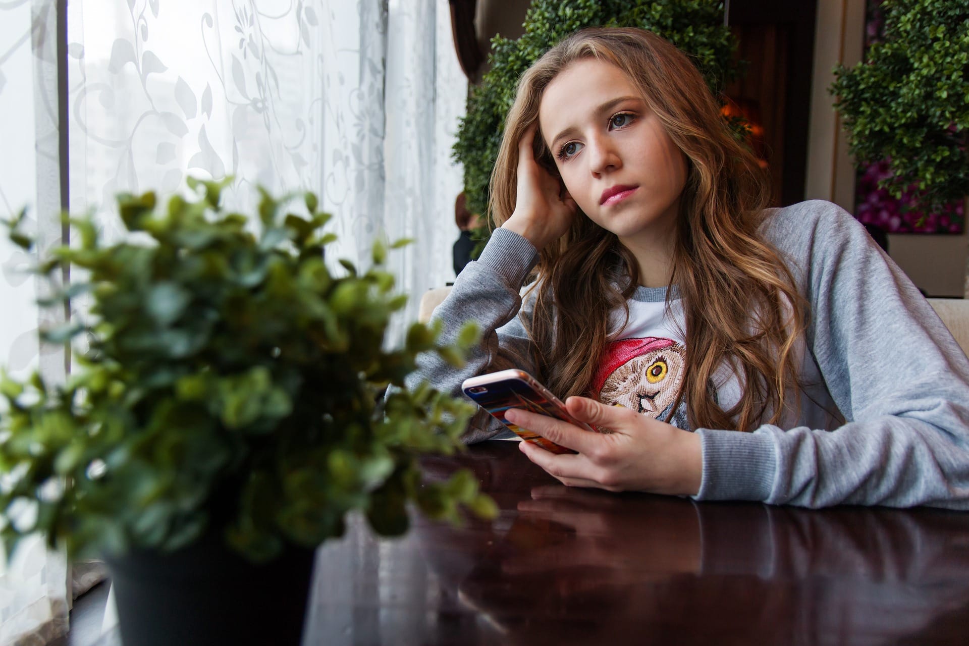 Ung jente sitter ved et bord og tenker samtidig som hun har mobilen oppe. 