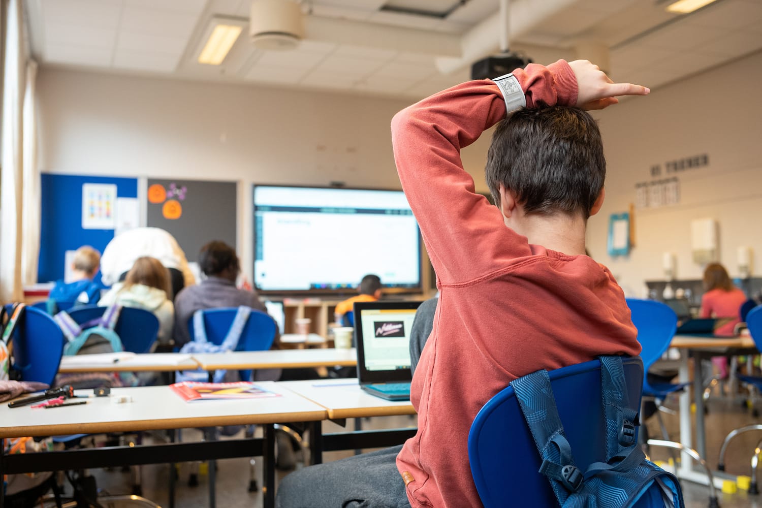 En gutt i rustrød genser sitter i klasserommet og rekker opp hånda