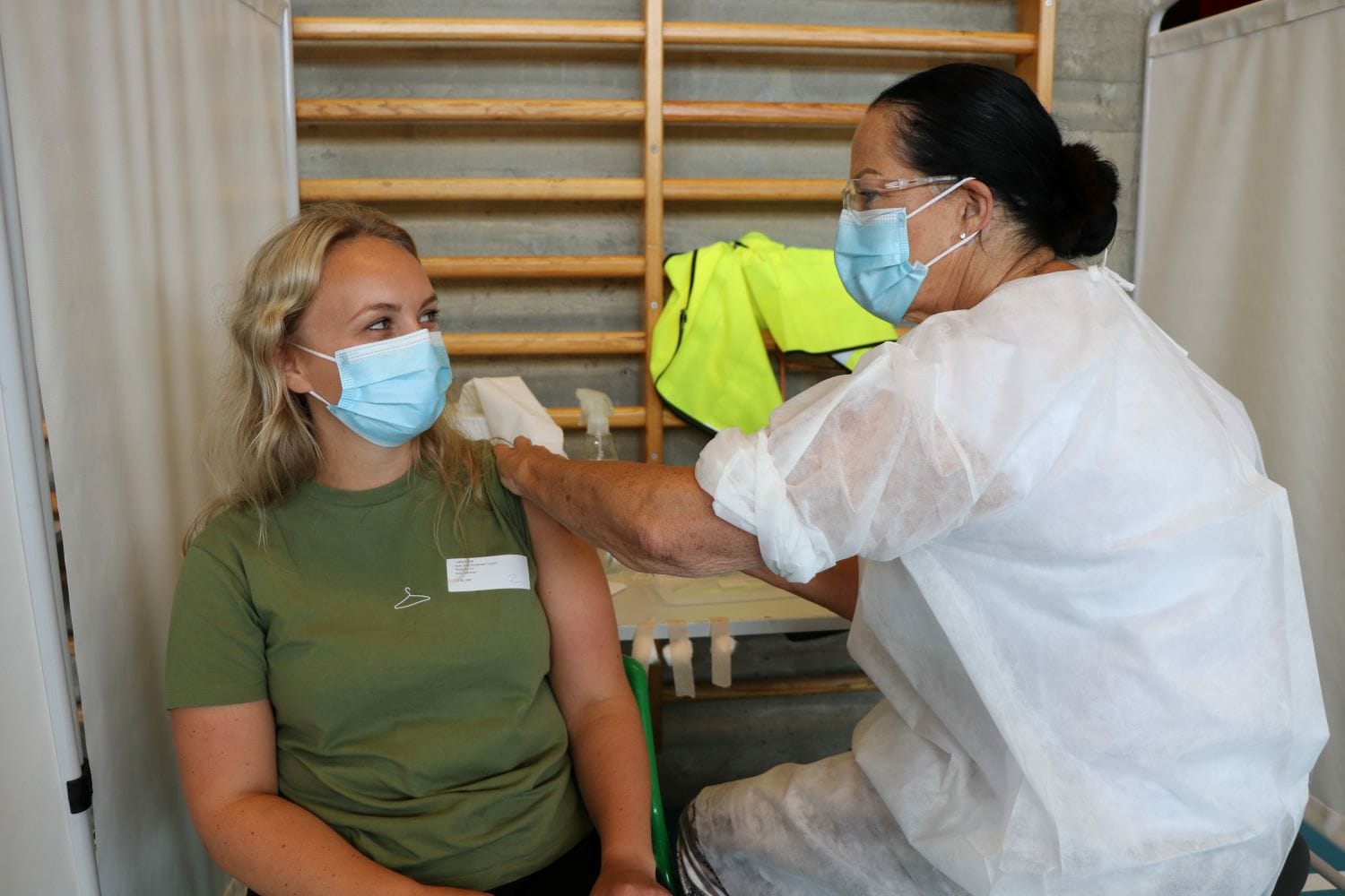 En lærer sitter i grønn trøye. Hun blir vaksinert av en sykepleier.