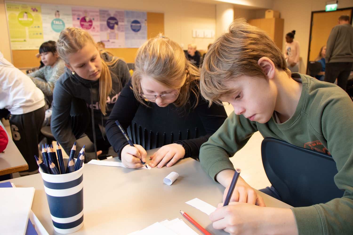 Tre elever, en gutt og to jenter, står bøyd over pulten og skriver på et ark.