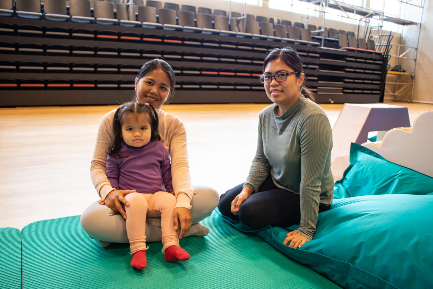 To kvinner og et barn på fanget til venstre sitter på gulvet på en grønn matte i gymsalen. 