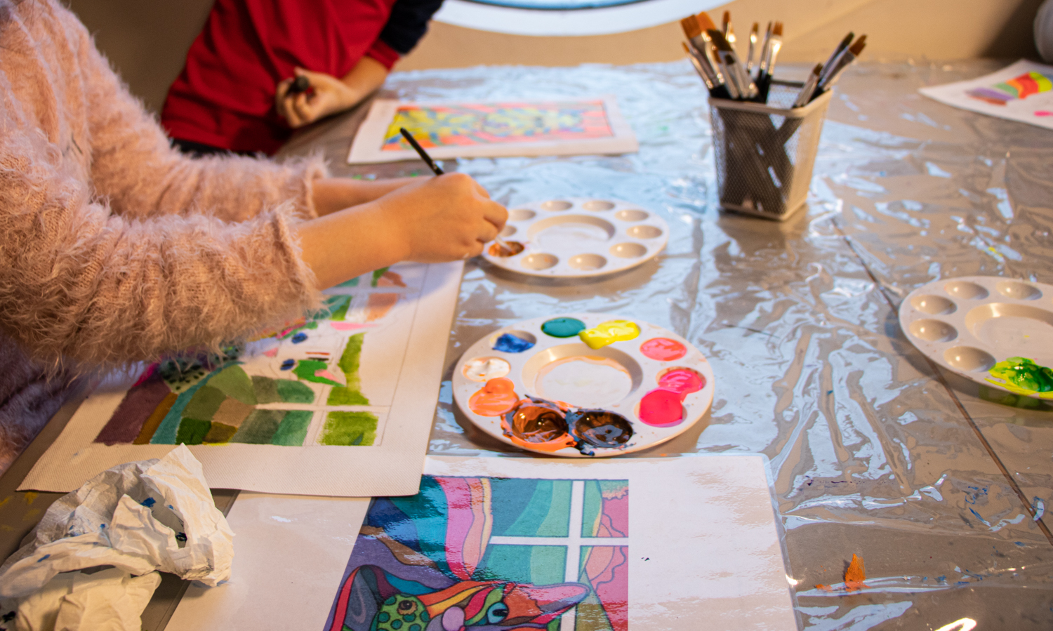Bord med maling i ulike farger, pensler og hendene til to barn som maler bilder.