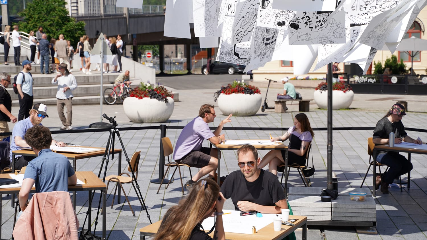 Flere innbyggere på Strømsø torg som sitter to og to overfor hverandre ved et bord. Over henger tegninger på hvite ark.