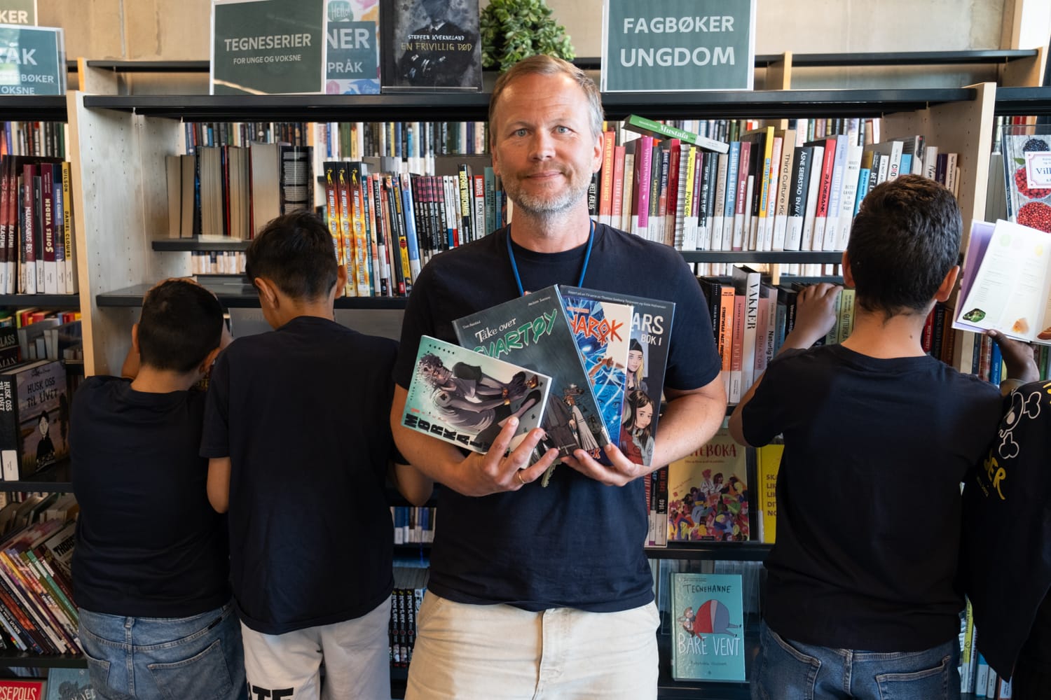 Skolebibliotekar Jørgen Gromstad viser hvilke bøker gutta har lest. Bak ham står gutta med ryggen til kameraet og leter etter nye bøker i bokhylla på biblioteket.