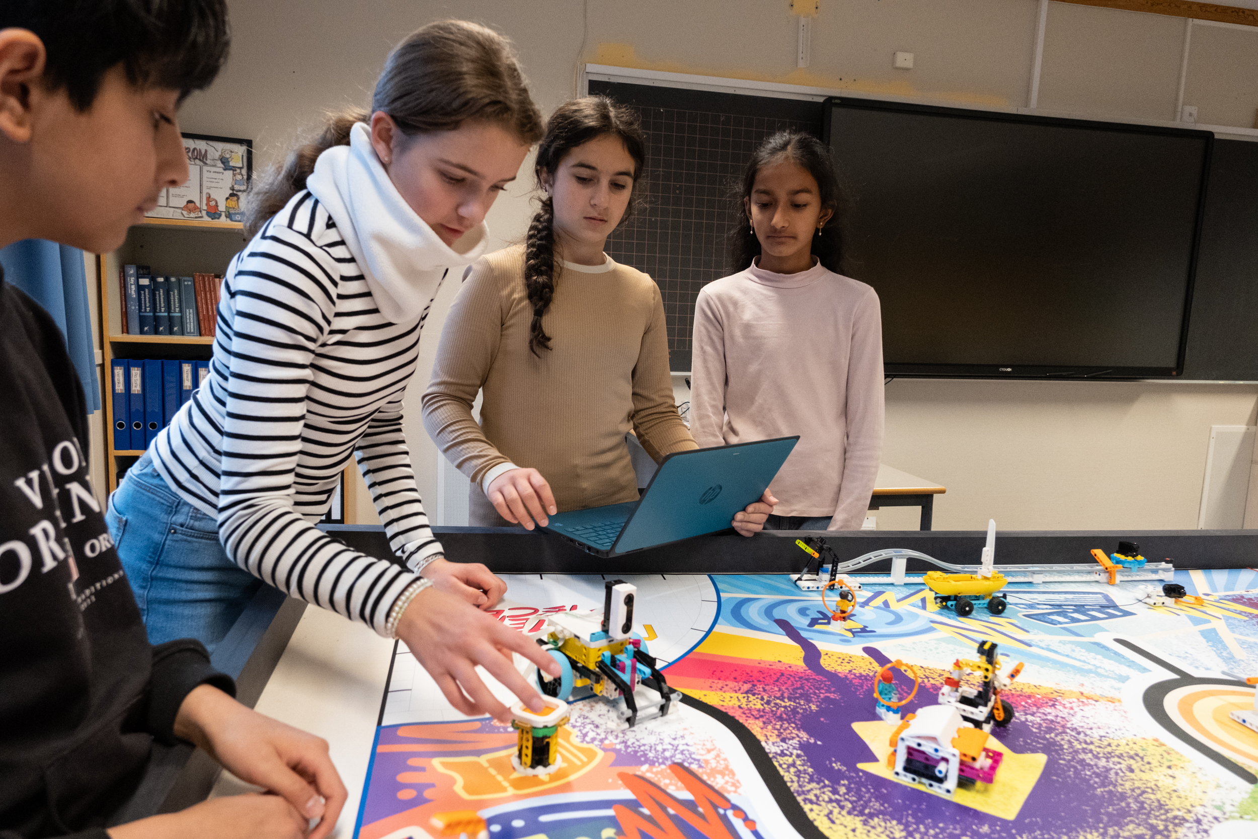 Fire elever rundt et fargerikt bord med legofigurer. En gutt i venstre hjørne og tre jenter som følger med på en robot på bordet i et klasserom.