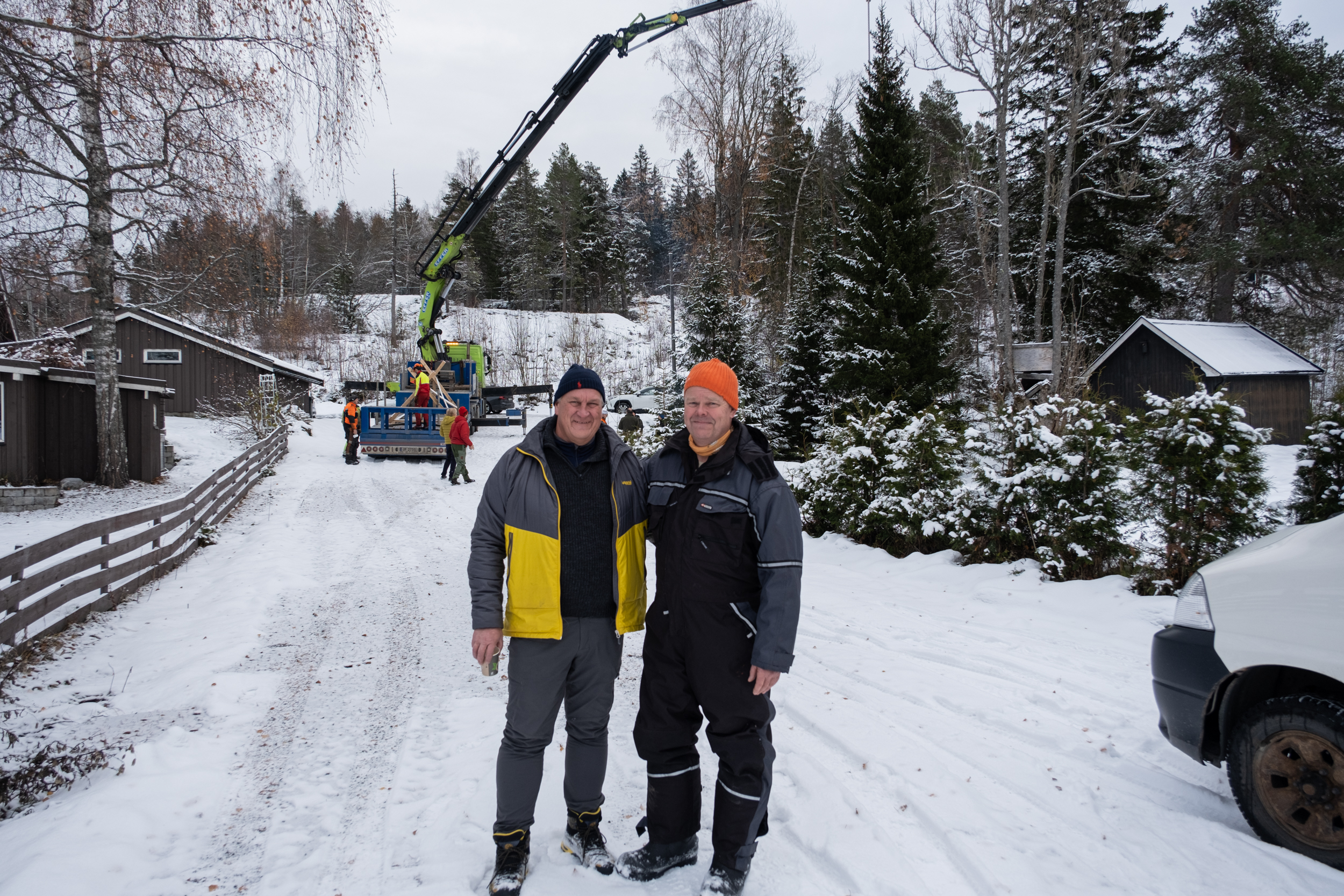 To menn står ute på vei med snø og smiler til kamera. I bakgrunn løftes et grantre med en stor kranbil.