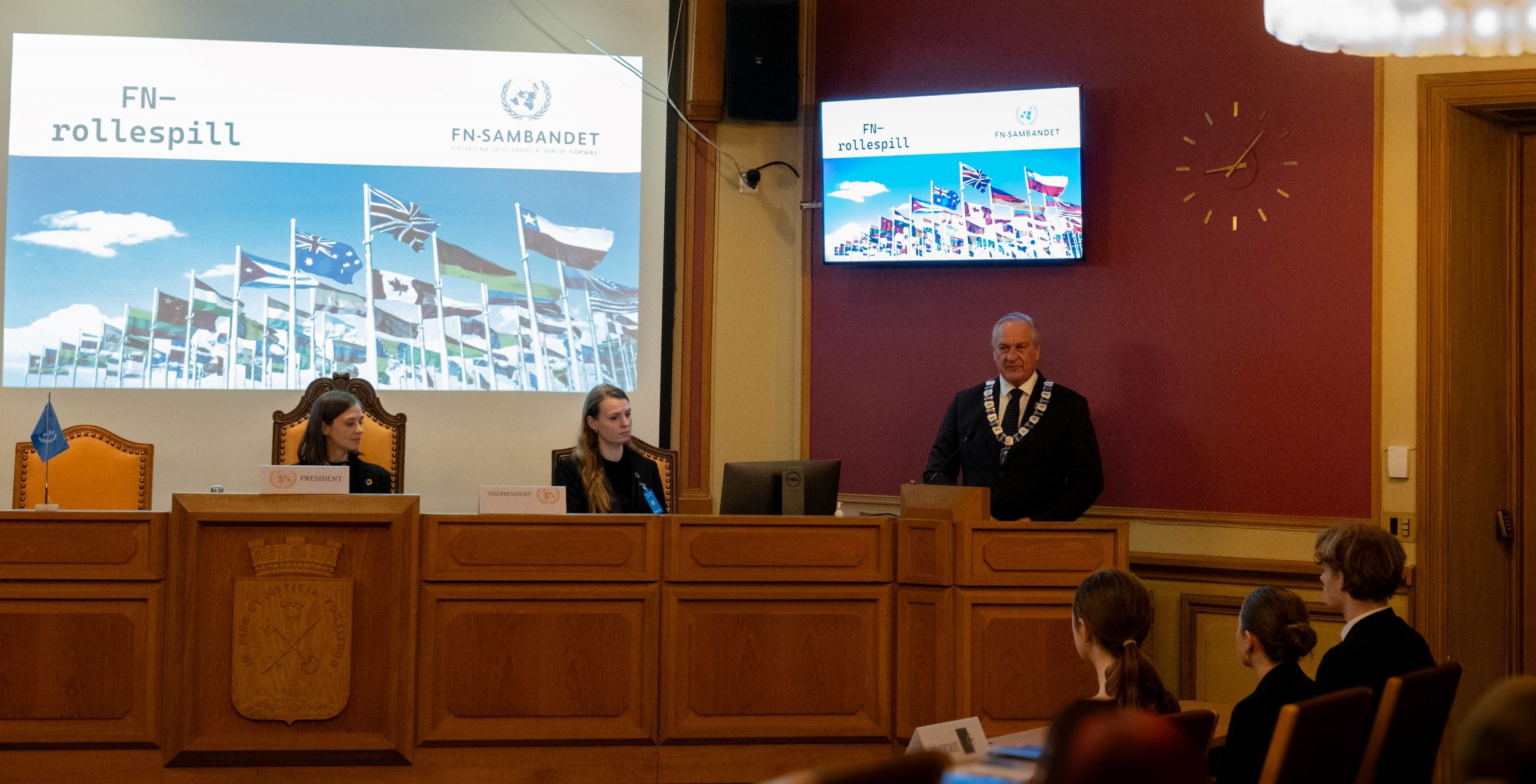 Siri Nikolaisen og Madeleine Hesstvedt Solstad fra FN-sambandet sitter på ordstyrerplann i kommunestyresalen i rådhuset i Drammen. Ordfører Kjell Arne Hermansen står på talerstolen og taler til salen.