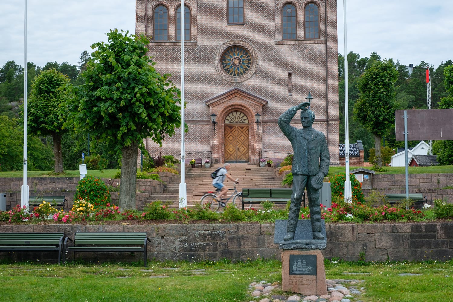 Kirkeparken i Svelvik sentrum. Svelvik kirke i bakgrunnen,  Statue og sitteplasser, samt beplantning.